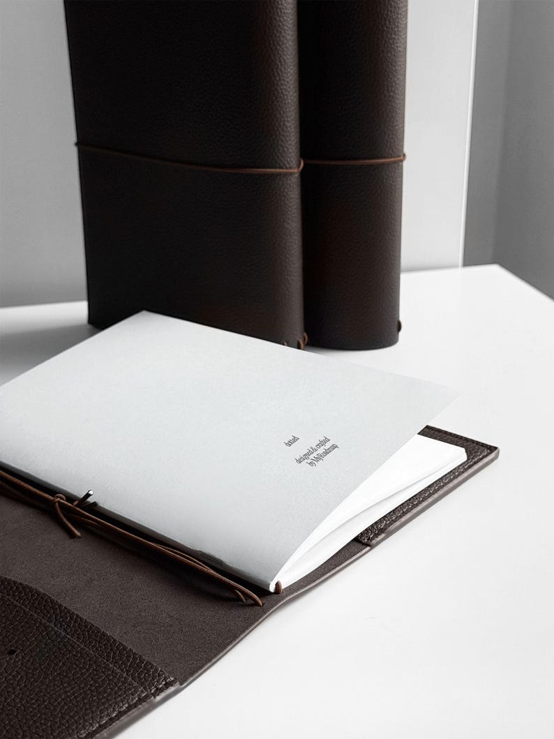 K&ouml;nnte beinhalten: Ein braunes Lederjournal mit passendem Lederbezug, ge&ouml;ffnet, um leere wei&szlig;e Seiten freizugeben. Zwei weitere Journale stehen im Hintergrund aufrecht. Der Text "brial designed & crafted by Mjolkman" ist auf der Seite aufgedruckt.