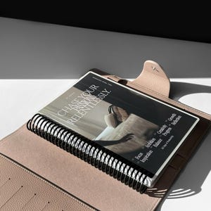 May include: A light pink leather bound journal with a black spiral binding. The journal is open to a page with the text "CHASE YOUR PASSIONS RELENTLESSLY." and a list of bullet points with the words "Focus, Ambition, Creativity, Growth, Inspiration, Balance, Progress, Reflection".