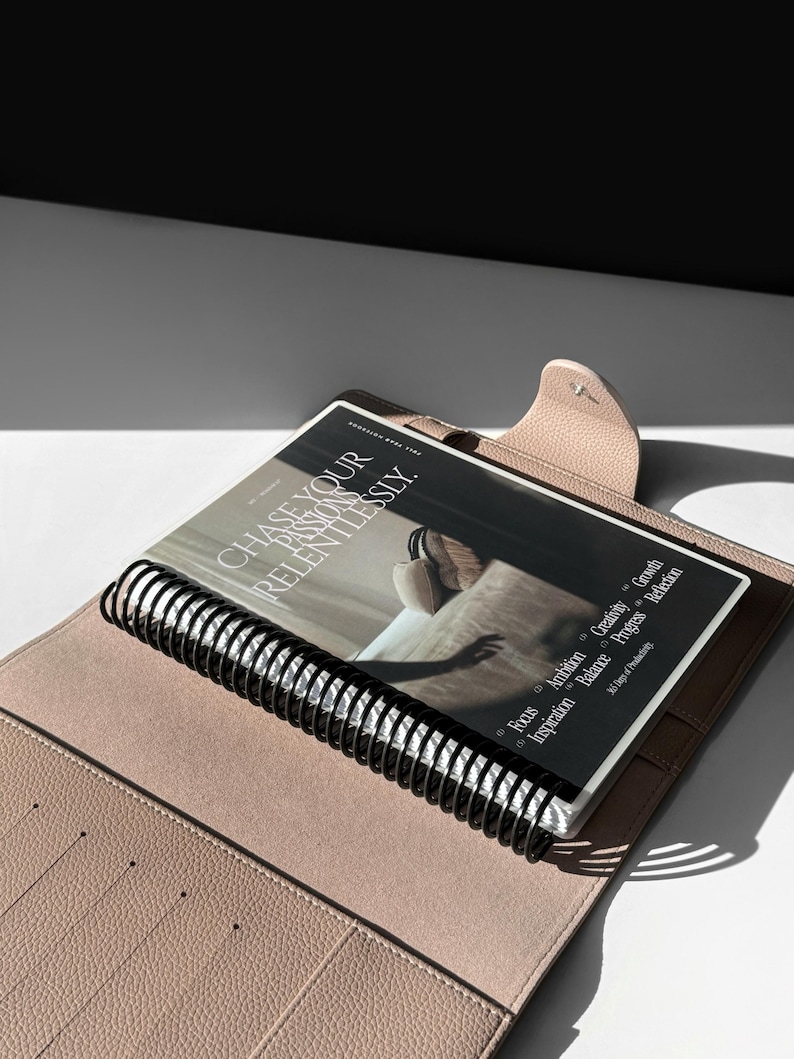 May include: A light pink leather bound journal with a black spiral binding. The journal is open to a page with the text "CHASE YOUR PASSIONS RELENTLESSLY." and a list of bullet points with the words "Focus, Ambition, Creativity, Growth, Inspiration, Balance, Progress, Reflection".
