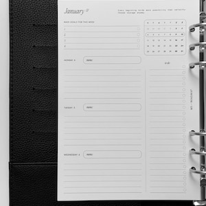May include: A black leather-textured binder containing a white planner insert. The page features a January calendar, a section for main goals, and daily planning fields for Monday, Tuesday, and Wednesday with memo boxes and a to-do list column.