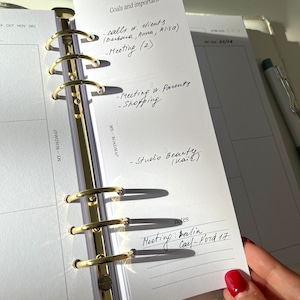 May include: A planner with gold-colored rings and white pages. The planner is open to a page with handwritten notes, including "Goals and important" and a list of tasks. A pen rests on the right side of the planner.