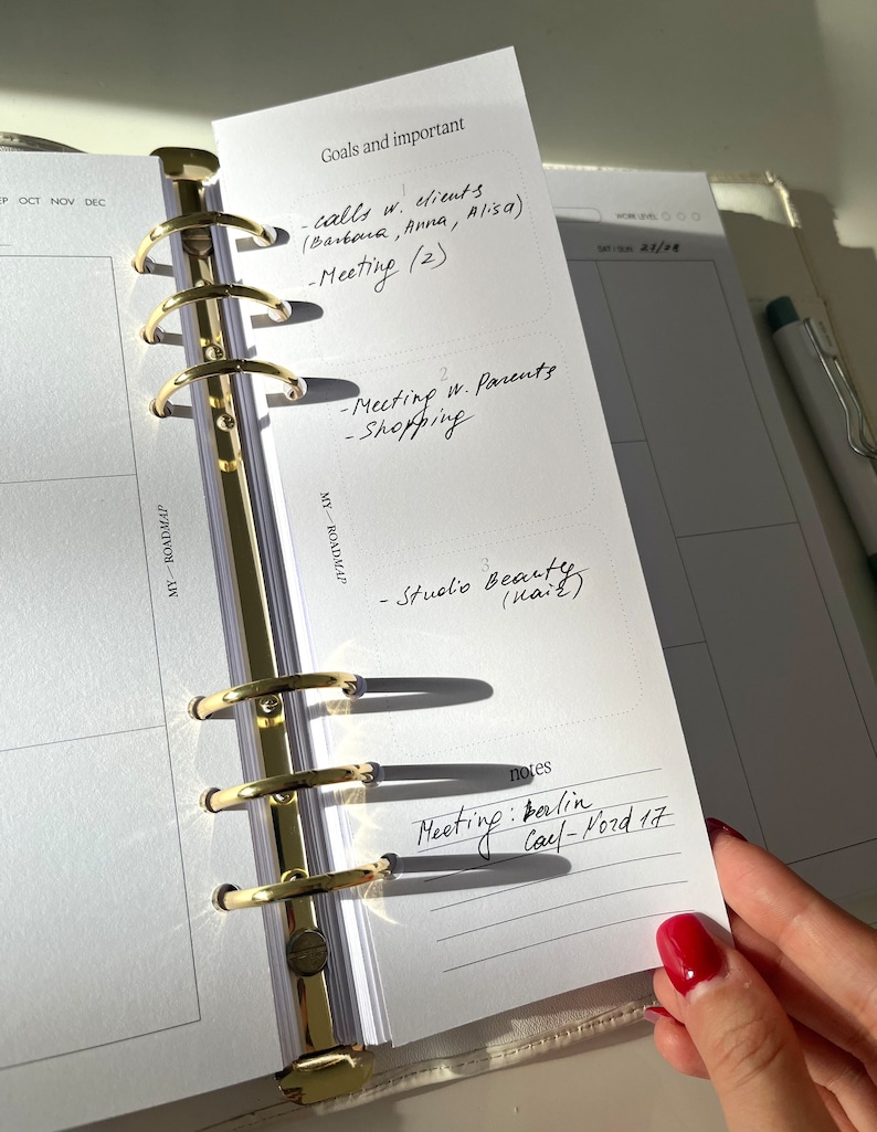 May include: A planner with gold-colored rings and white pages. The planner is open to a page with handwritten notes, including "Goals and important" and a list of tasks. A pen rests on the right side of the planner.