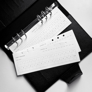 Può includere: Un'agenda rilegata in pelle nera con inserti di tracciamento delle abitudini bianchi. Gli inserti sono etichettati "Habit Tracker" e hanno una griglia di numeri per ogni giorno del mese. L'agenda ha un sistema di rilegatura ad anelli.