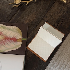 K&ouml;nnte beinhalten: Ein offenes Notizbuch mit kariertem Papier und einem braunen Ledercover liegt auf einer dunklen Holzoberfl&auml;che. Ein Buch mit einer Tulpenillustration und ein goldfarbener Ast mit Kerzenhaltern sind ebenfalls zu sehen.