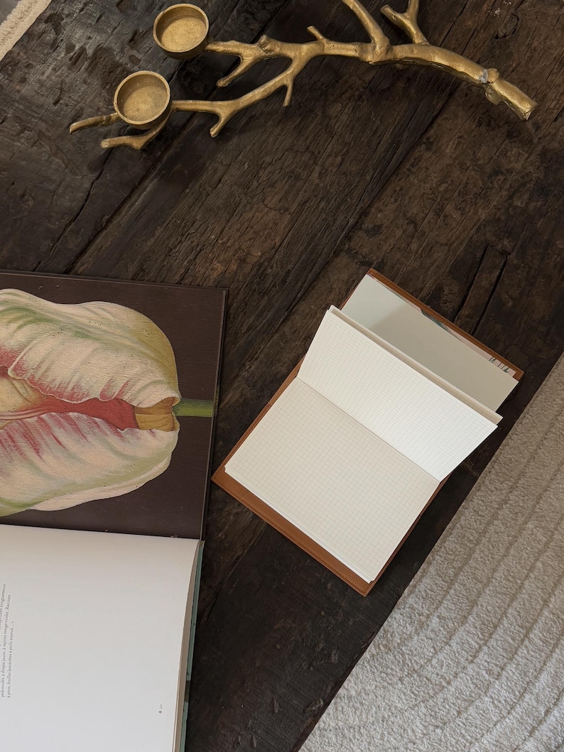 K&ouml;nnte beinhalten: Ein offenes Notizbuch mit kariertem Papier und einem braunen Ledercover liegt auf einer dunklen Holzoberfl&auml;che. Ein Buch mit einer Tulpenillustration und ein goldfarbener Ast mit Kerzenhaltern sind ebenfalls zu sehen.