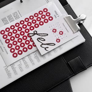 May include: A black leather-textured planner containing white pages with printed text, including the words "Focus," "Balance," and "Flexibility." A sheet of red circular stickers and a small calendar card labeled "Feb" are attached with a silver metal clip.