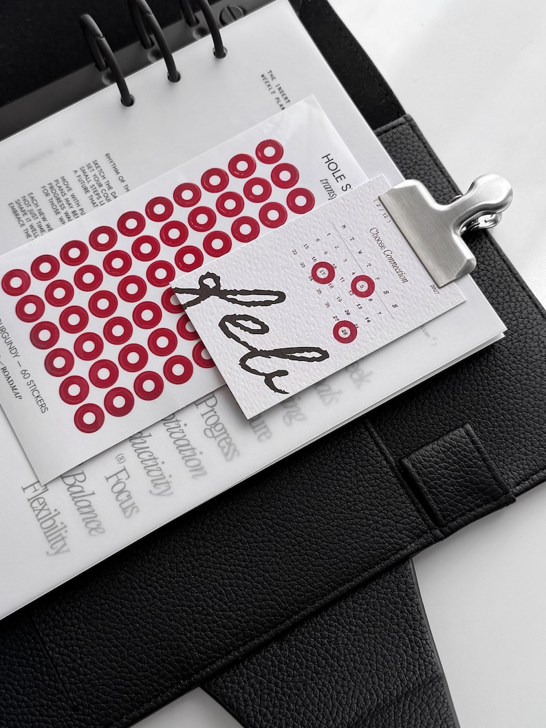 May include: A black leather-textured planner containing white pages with printed text, including the words "Focus," "Balance," and "Flexibility." A sheet of red circular stickers and a small calendar card labeled "Feb" are attached with a silver metal clip.