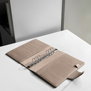 May include: A beige leather bound planner with a silver ring binder and a flap closure.