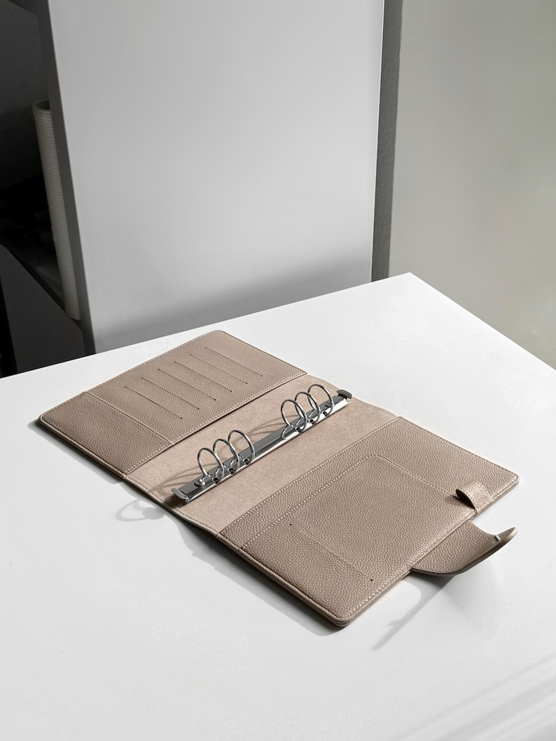 May include: A beige leather bound planner with a silver ring binder and a flap closure.