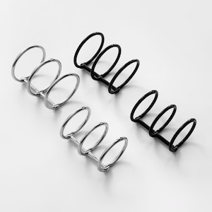 Könnte beinhalten: Vier Ordnerringe, zwei in Silber und zwei in Schwarz, sind auf einer weißen Oberfläche angeordnet. Jedes Ring-Set hat drei kreisförmige Ringe, die an der Basis verbunden sind, was ein funktionales und minimalistisches Design ergibt.