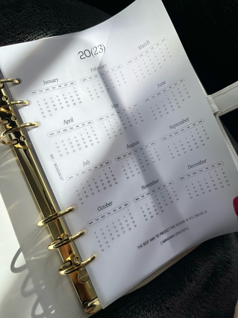 Pu&ograve; includere: Una pagina di agenda bianca con un calendario 2023 da gennaio a dicembre, anelli di rilegatura in metallo dorato sul lato sinistro, il testo MY-ROADMAP e la citazione THE BEST WAY TO PREDICT THE FUTURE IS TO CREATE IT (ABRAHAM LINCOLN).
