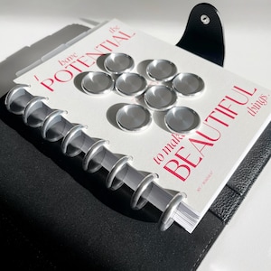 Może przedstawiać: Czarny skórzany planer z srebrnymi kółkami i otwartą białą stroną na stronie z tekstem "Mam potencjał, aby tworzyć piękne rzeczy." Strona ma srebrne naklejki w kształcie dysków.