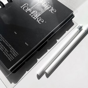 May include: A black planner with a white cover that says "No time for fake" and a white background. The planner is open to a page with the words "Too Busy" and a quote about the importance of genuine progress. Two white styluses are lying on the white surface.