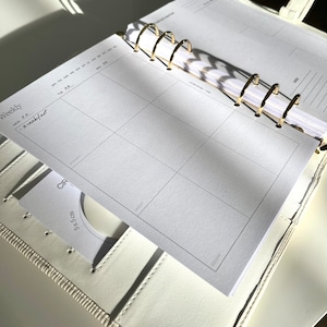 May include: Open white leather planner with gold-colored rings, featuring a weekly layout. The planner is open to a page with a grid for daily scheduling. A small card with a 5x5 cm measurement is visible. The word "Weekly" is printed on the page.