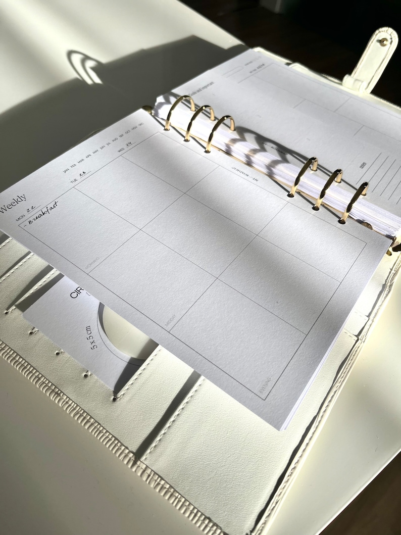 May include: Open white leather planner with gold-colored rings, featuring a weekly layout. The planner is open to a page with a grid for daily scheduling. A small card with a 5x5 cm measurement is visible. The word "Weekly" is printed on the page.