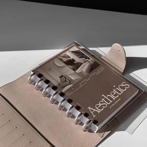 May include: A beige leather bound planner with a clear plastic page protector holding a book titled "Aesthetics in the Everyday". The book has a brown background with a black and white photo of a room.