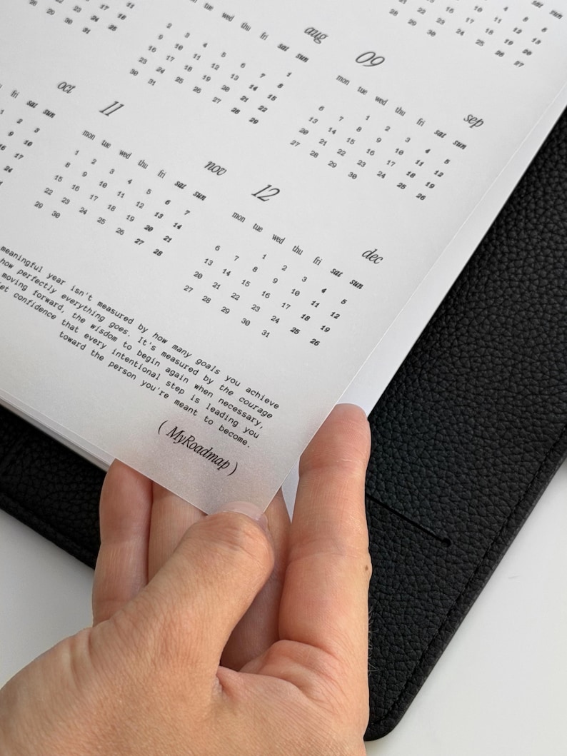 May include: A black leather planner containing a white calendar insert with months from August to December. The page features a printed quote about personal growth and the text MyRoadmap in parentheses at the bottom of the translucent sheet.