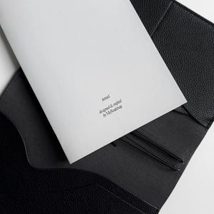 K&ouml;nnte beinhalten: Eine schwarze Leder-Journalh&uuml;lle mit einem wei&szlig;en Notizblock im Inneren. Auf dem Notizblock steht das Wort "dotted" und der Text "designed & crafted by MyRoadmap". Zwei schwarze Gummib&auml;nder sind sichtbar.