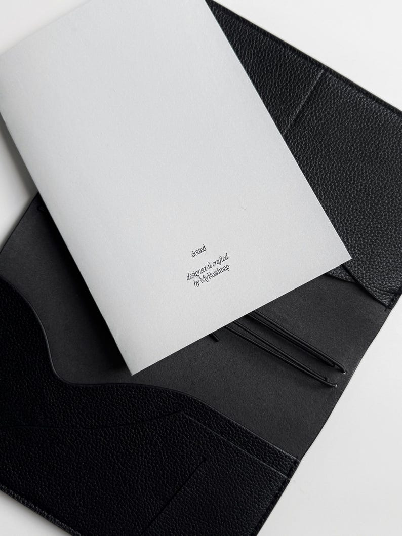 K&ouml;nnte beinhalten: Eine schwarze Leder-Journalh&uuml;lle mit einem wei&szlig;en Notizblock im Inneren. Auf dem Notizblock steht das Wort "dotted" und der Text "designed & crafted by MyRoadmap". Zwei schwarze Gummib&auml;nder sind sichtbar.