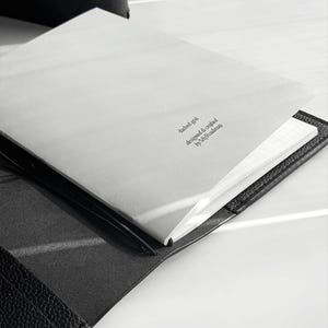May include: Open black leather journal with a white, lined paper interior. The top page has the text "dashed grid designed & crafted by MyRoadmap" printed in a small, dark font. The journal is open on a white surface.
