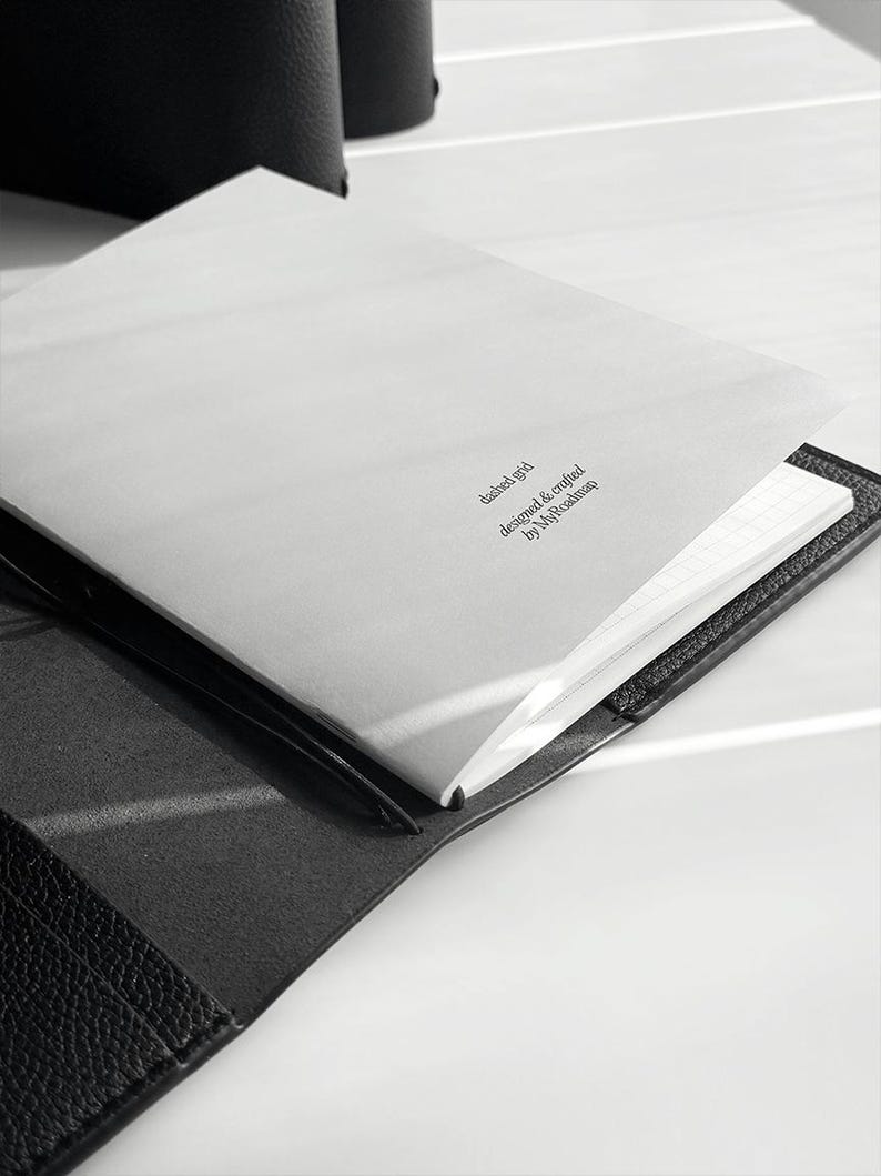 May include: Open black leather journal with a white, lined paper interior. The top page has the text "dashed grid designed & crafted by MyRoadmap" printed in a small, dark font. The journal is open on a white surface.