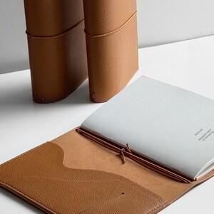 K&ouml;nnte beinhalten: Drei braune Lederjournale. Eines ist ge&ouml;ffnet und zeigt wei&szlig;es Papier. Das offene Journal hat eine Tasche auf der Innenseite des Einbands. Die Journale sind mit braunem Faden gebunden.
