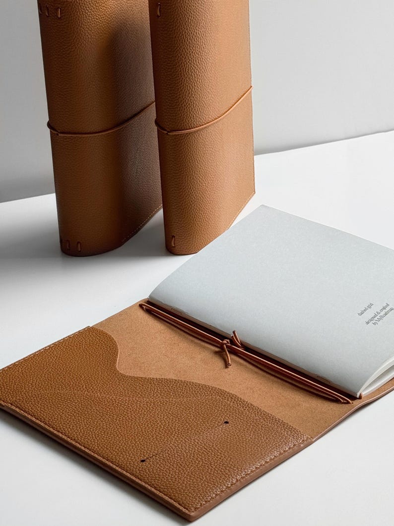 K&ouml;nnte beinhalten: Drei braune Lederjournale. Eines ist ge&ouml;ffnet und zeigt wei&szlig;es Papier. Das offene Journal hat eine Tasche auf der Innenseite des Einbands. Die Journale sind mit braunem Faden gebunden.