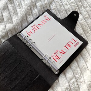 Może przedstawiać: Czarny sk&oacute;rzany segregator z białym wkładem. Wkładka zawiera czerwony tekst o treści "I have the POTENTIAL to make BEAUTIFUL things." Segregator jest otwarty, ukazując srebrne pierścienie i wewnętrzne kieszenie. Tło to biała, teksturowana powierzchnia.