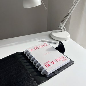 Może przedstawiać: Czarny sk&oacute;rzany planer z białym notatnikiem z napisem "bare the POTENTIAL to make BEAUTIFUL things". Widoczna jest r&oacute;wnież biała lampa biurkowa i długopis. Planer jest otwarty, ukazując strony notatnika.