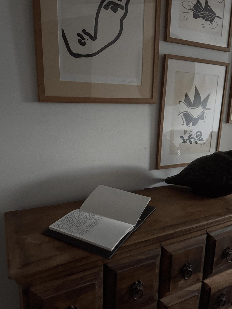 May include: An open notebook with handwritten text rests on a dark wooden dresser. Above, framed art pieces in neutral tones hang on a white wall. The artwork includes abstract line drawings and shapes, creating a minimalist aesthetic.