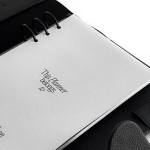 May include: Black leather planner with silver metal rings and a white page that reads "This Planner belongs to". The planner is open, revealing the interior pages and the textured leather cover.