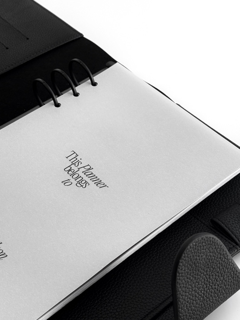 May include: Black leather planner with silver metal rings and a white page that reads "This Planner belongs to". The planner is open, revealing the interior pages and the textured leather cover.