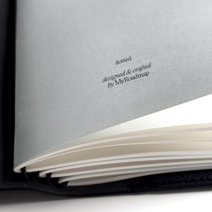 K&ouml;nnte beinhalten: Ein teilweise ge&ouml;ffnetes schwarzes Notizbuch mit wei&szlig;en, gepunkteten Seiten. Der Text "dotted" und "designed & crafted by MyRoadmap" ist auf einer grauen Seite aufgedruckt. Das Notizbuch ist ge&ouml;ffnet.