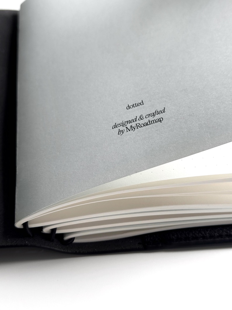 K&ouml;nnte beinhalten: Ein teilweise ge&ouml;ffnetes schwarzes Notizbuch mit wei&szlig;en, gepunkteten Seiten. Der Text "dotted" und "designed & crafted by MyRoadmap" ist auf einer grauen Seite aufgedruckt. Das Notizbuch ist ge&ouml;ffnet.
