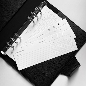 Può includere: Un'agenda rilegata in pelle nera con un anello di rilegatura argentato. L'agenda contiene diverse pagine bianche con griglie di tracciamento delle abitudini e il testo "HABIT TRACKER" e "HABIT NAME".
