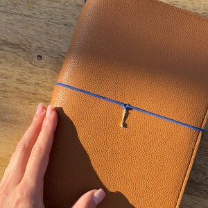 K&ouml;nnte beinhalten: Ein hellbraunes Lederjournal mit strukturierter Oberfl&auml;che. Das Journal wird mit einem blauen Gummiband und einem goldfarbenen Verschluss gesichert. Das Journal ist rechteckig und scheint geschlossen zu sein.