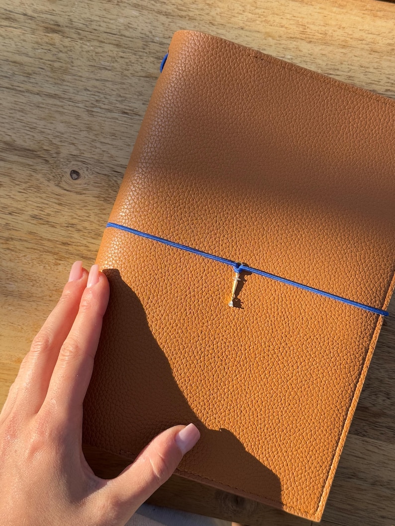 K&ouml;nnte beinhalten: Ein hellbraunes Lederjournal mit strukturierter Oberfl&auml;che. Das Journal wird mit einem blauen Gummiband und einem goldfarbenen Verschluss gesichert. Das Journal ist rechteckig und scheint geschlossen zu sein.