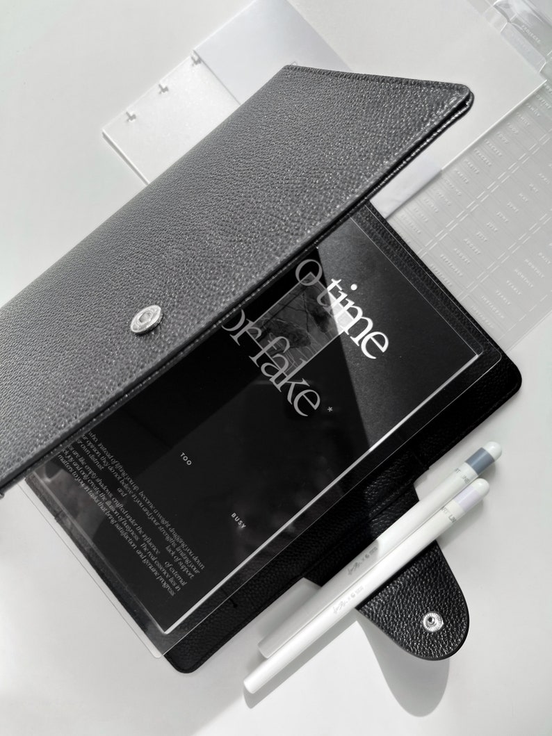 May include: A black leather planner with a clear plastic cover. The cover has the text "time or fake" printed on it. The planner is open to a page with the text "too busy" printed on it. Two white pens are lying on top of the planner.