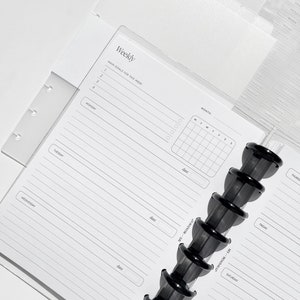 May include: A white weekly planner with a black coil binding. The planner has a section for main goals for the week, a monthly calendar, and a daily planner for each day of the week. The planner is designed for use with a disc-bound system.