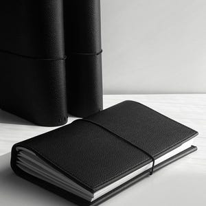 May include: A black leather journal with a textured cover and elastic closure, lying open on a white surface. Two additional journals stand upright in the background, also black with elastic closures. The journals have white-edged pages.