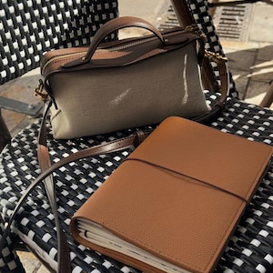 K&ouml;nnte beinhalten: Eine beigefarbene Canvas-Handtasche mit braunen Lederbes&auml;tzen und einem passenden braunen Ledergriff und -riemen. Ein braunes Lederjournal mit Riemenverschluss liegt auf einem schwarz-wei&szlig; gewebten Stuhl.