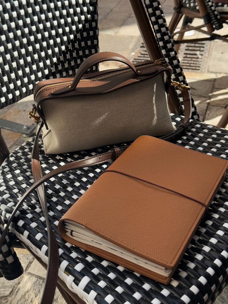 K&ouml;nnte beinhalten: Eine beigefarbene Canvas-Handtasche mit braunen Lederbes&auml;tzen und einem passenden braunen Ledergriff und -riemen. Ein braunes Lederjournal mit Riemenverschluss liegt auf einem schwarz-wei&szlig; gewebten Stuhl.