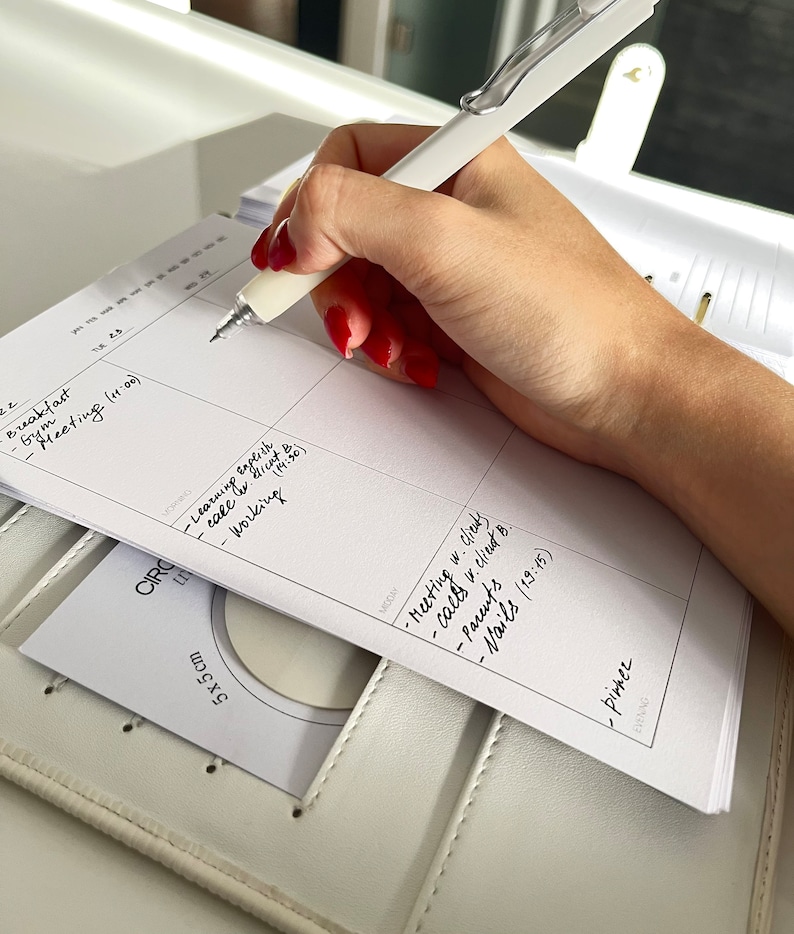 May include: A white pen writing on a planner with handwritten notes. The planner is open to a weekly schedule with the text "Breakfast - Gym - Meeting (11:00)". A small card with "5x5 cm" is visible.