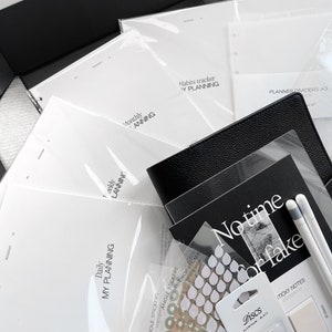 May include: A black box filled with planner accessories, including a black leather planner, white paper inserts, stickers, and a black pen. The inserts include a daily, weekly, monthly, and habits tracker. The box also includes a pack of black and white sticky notes.