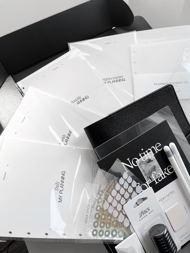May include: A black box filled with planner accessories, including a black leather planner, white paper inserts, stickers, and a black pen. The inserts include a daily, weekly, monthly, and habits tracker. The box also includes a pack of black and white sticky notes.