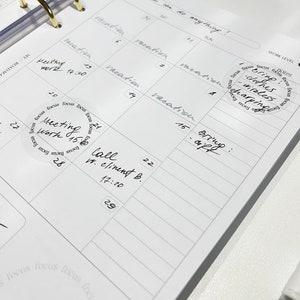 May include: A white planner with a gold ring binder. The planner has a weekly layout with dates and days of the week labeled. There are several handwritten notes and stickers on the planner, including a sticker that says "Bring clothes - wireless charging".
