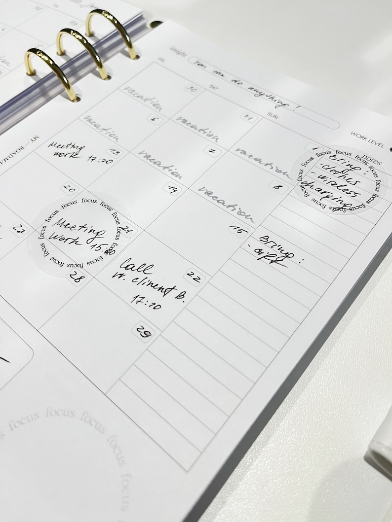 May include: A white planner with a gold ring binder. The planner has a weekly layout with dates and days of the week labeled. There are several handwritten notes and stickers on the planner, including a sticker that says "Bring clothes - wireless charging".