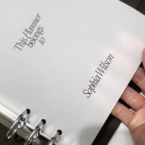 May include: A white planner page with the text "This Planner belongs to" and "Sophia Wilson" in elegant script. The page is held open in a dark brown binder with silver rings.