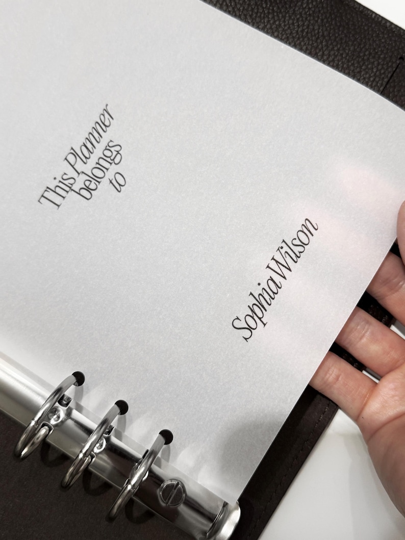 May include: A white planner page with the text "This Planner belongs to" and "Sophia Wilson" in elegant script. The page is held open in a dark brown binder with silver rings.