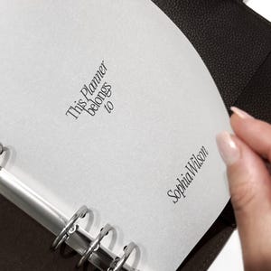 May include: A dark brown leather planner with silver metal rings. The open page reads "This Planner belongs to" and "Sophia Wilson" in elegant script. The planner is partially open, revealing the interior pages.